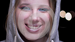 Sportsgirl in hoodie with colorful make-up raising her eyes shyly and laughing into camera on blurred lights background.