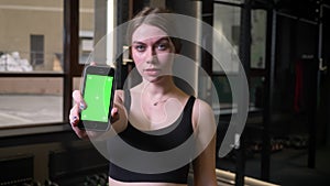 Sportsgirl showing smartphone with chromakey green screen into camera being serious and concentrated in gym.