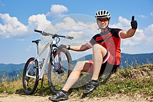 Sportsman bicyclist in sportswear and helmet sitting near his mountain bike on grassy roadside