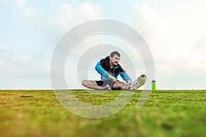 Sportsman with a bottle of water sitting and doing warming stretching exercise in the park