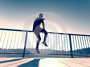 Sportsman doing stretching at handrail in park. Silhouette against sun,