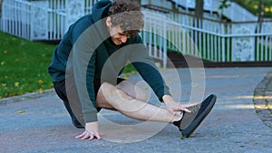 Sportsman sitting and stretching his knee, leg, muscles in green park. Young Caucasian man suffering from strong pain in