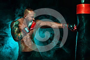 Sportsman with tattoos, man boxer fighting in gloves with boxing punching bag. Isolated on black background with smoke
