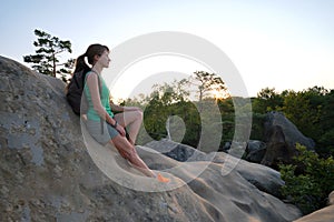 Sportswoman sitting alone taking a break on hillside trail. Female hiker enjoying view of evening nature from rocky