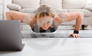 Sportswoman training at home. Muscular adult woman with fitness tracker doing push ups, near laptop in living room