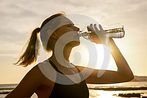 sporty woman drinking water from bottle after outdoor fitness workout at sunset