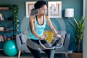 Sporty african young woman exercising on smart stationary bike and listening to music at home
