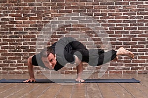 Sporty attractive young man working out, yoga, pilates, fitness training, asana Dwi pada kaundiniasana over brick wall background