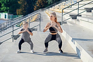 Sporty beautiful elder and younger sisters which squating together during fitness training outdoors.