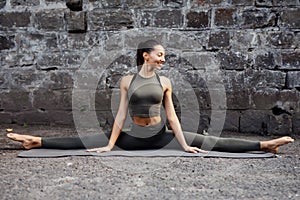 Sporty beautiful young woman practicing yoga hanumanasana working out, wearing sportswear, outdoor full length, brick wall backgro