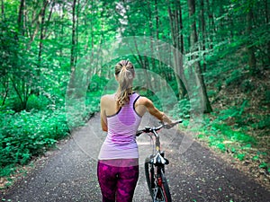 Sporty caucasian woman with blonde hair walking with her bike backward in a forest alley