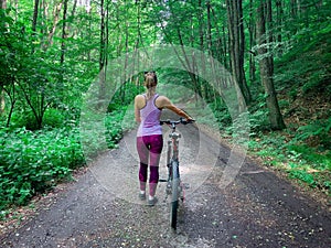 Sporty caucasian woman with blonde hair walking with her bike backward in a forest alley