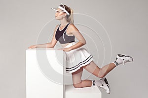 Sporty concept. Side view portrait of beautiful athletic young woman in fashionable sportwear standing on knee on white cube and