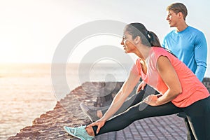 Sporty couple stretching legs in front of ocean at sunset - young people training and making legs exercises outdoor