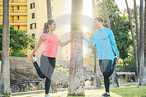 Sporty couple stretching legs leaning on a tree in park at sunset - Two persons training and making legs exercises outdoor