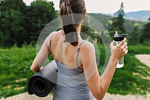 Sporty female holding yoga mat and bottle of water at sunny mountain hills. Healthy outdoor workout