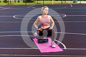 Sporty fit girl training on mat outdoor summer day, using foam roller massager to support knee while stretching legs