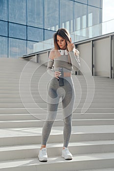 Sporty Fit Woman in a Tight Gray Tracksuit Poses with a Bottle of Water on the Stairs Outdoors