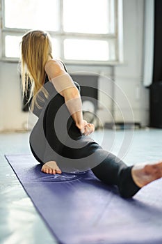 Sporty flexible blonde woman in black sportswear practicing yoga in yoga class, making stretching back on mat.