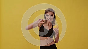 Sporty girl connecting headphones to orange and listens to music. Studio, yellow background.