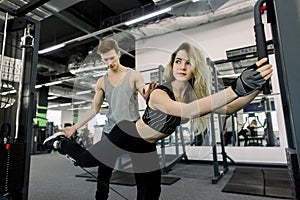 Sporty girl doing stratching legs exercises with assistance of her personal trainer at public gym. Coaching assistance