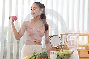 Sporty girl in fitness sport wear lifting dumbbell, fruit vegetable and healthy ingredients on wooden table at kitchen. Healthy