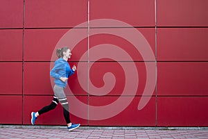 Sporty girl runs against a red wall in the street, a woman jogging in sportswear outdoors, copy space