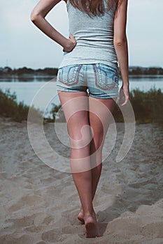 Sporty girl spending time by a lake back-view. Rear view of young woman relaxing outside wearied denim shorts and tank