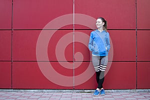 Sporty girl stands against a red wall in sportswear, fitness woman posing on a city street, copy space