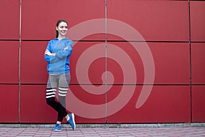 Sporty girl stands against a red wall in sportswear, fitness woman posing on a city street, copy space