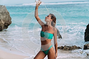 Sporty girl in swimsuit at beach pours clear water on herself from bottle