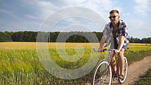 Sporty guy in sunglasses cycling along country trail outdoor. Young smiling man riding vintage bicycle at rural road
