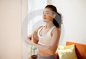 Sporty lady meditating with hands clasped standing in living room