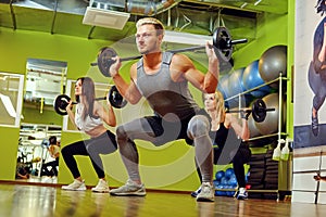 Sporty male and two females doing squats with barbells in a gym.