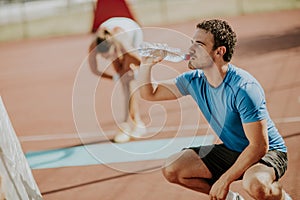Sporty man drinking water while young woman doing exercise in th
