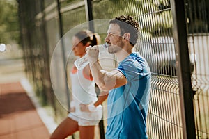 Sporty man drinking water while young woman doing exercise in th