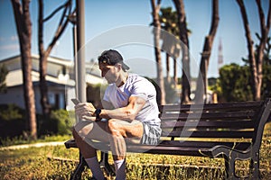 Sporty man personal trainer sitting looking at his phone on a bench in an outdoor park. Muscle body and well defined. Sporty