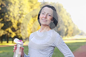 Sporty mature woman hold bottle with water outdoor on sunny day in the park