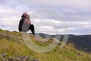 Sporty mature woman sits on top of a mountain and looks through binoculars at the Ural village, at the mountains, at the taiga, at