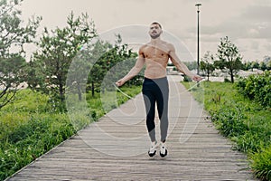 Sporty muscular man uses skipping rope for cardio training outdoor, has perfect body, breathes fresh air and demonstrates