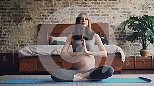 Sporty pregnant woman is practising yoga at home sitting on mat on bedroom floor raising hands, putting palms together