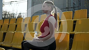 Sporty pretty female sits at staduim tribune, drinks water from plastic bottle