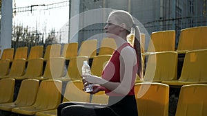 Sporty pretty female sits at staduim tribune, drinks water from plastic bottle