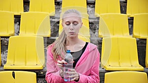 Sporty pretty female sits at staduim tribune, drinks water from plastic bottle