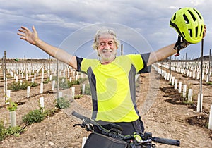 Sporty retired bearded man with bicycle in countryside smiling happy with raised arms and helmet in the hand - elderly people with