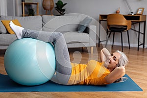 Sporty senior lady exercising her abs muscles with fitball on yoga mat at home, looking and smiling at camera