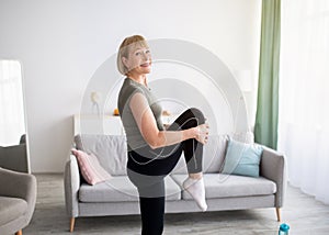 Sporty senior woman stretching her leg and smiling at camera indoors, free space. Domestic trainings during coronavirus