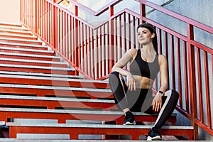 Sporty slim happy young beautiful woman in black fashionable sportwear sitting on the red stairs and relaxing during outdoor