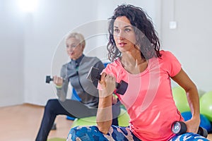 Sporty slim women taking part in gym fitness class exercising sitting on physioballs doing alternated biceps curl with