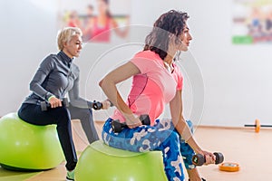 Sporty slim women taking part in gym fitness class exercising sitting on physioballs doing alternated biceps curl with